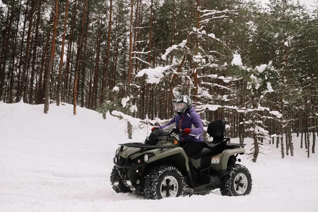 Förare med hjälm kör en fyrhjuling genom en snöig tallskog, en bild för ATV-försäkring