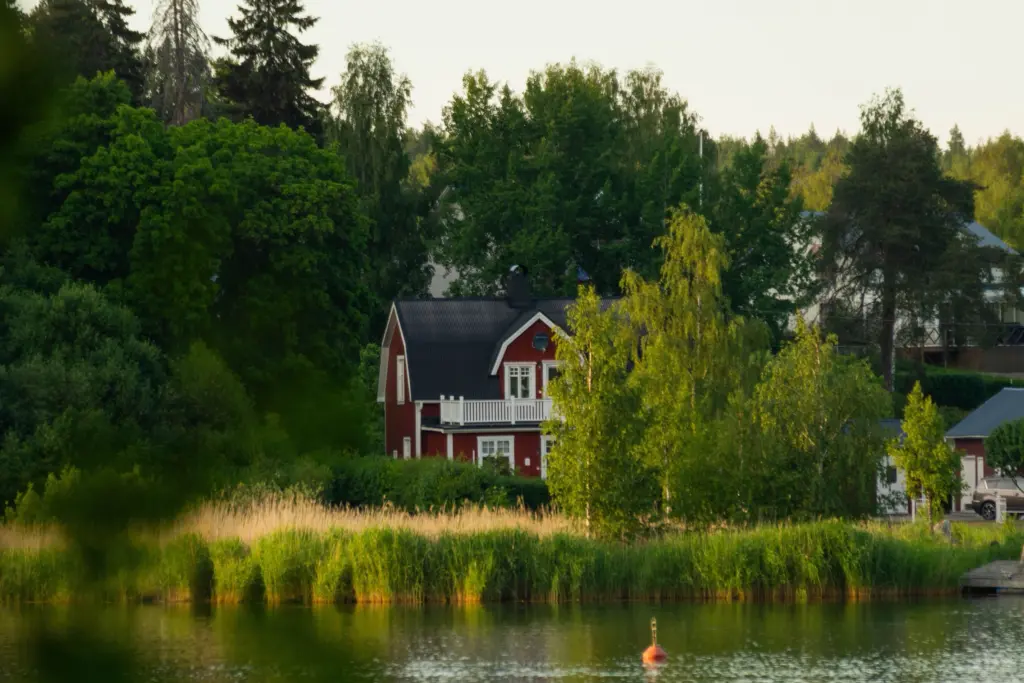 Rött trähus med vita knutar bland björkar och granar vid en lugn sjö med vass en sommarkväll