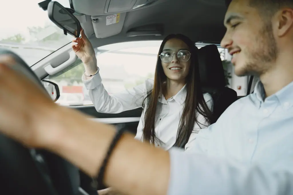 Ung förare ställer in backspegeln med en vän bredvid sig i bilen, unga förare betalar ofta mer för bilförsäkringen