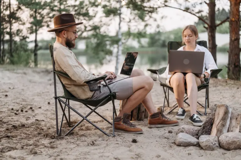 Två personer jobbar på sina datorer i campingstolar vid en sjö bland tallar, i sommarstugan behövs bredband bara några månader om året