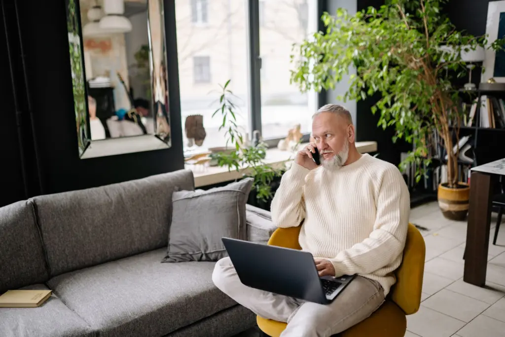Man i stickad tröja sitter i en fåtölj i vardagsrummet med en laptop i knät och pratar i telefon, som när man ringer sin bredbands- eller tv-leverantör