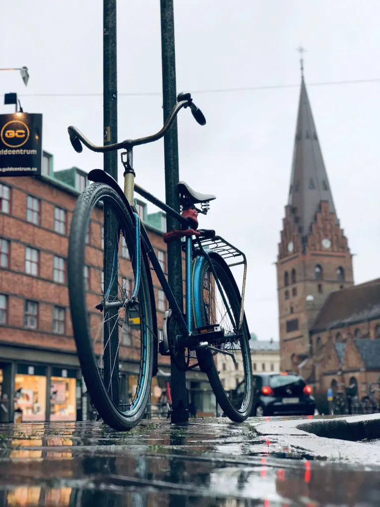 Cykel fastlåst vid en stolpe på en regnvåt gata i Malmö med en kyrka i tegel i bakgrunden, en låst cykel är ett krav för ersättning vid cykelstöld