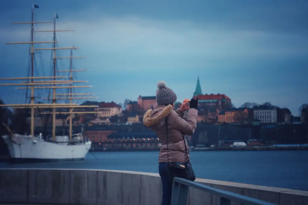 Kvinna i vinterjacka och mössa fotograferar med mobilen vid vattnet i Stockholm, med ett segelfartyg och stadens hus i bakgrunden