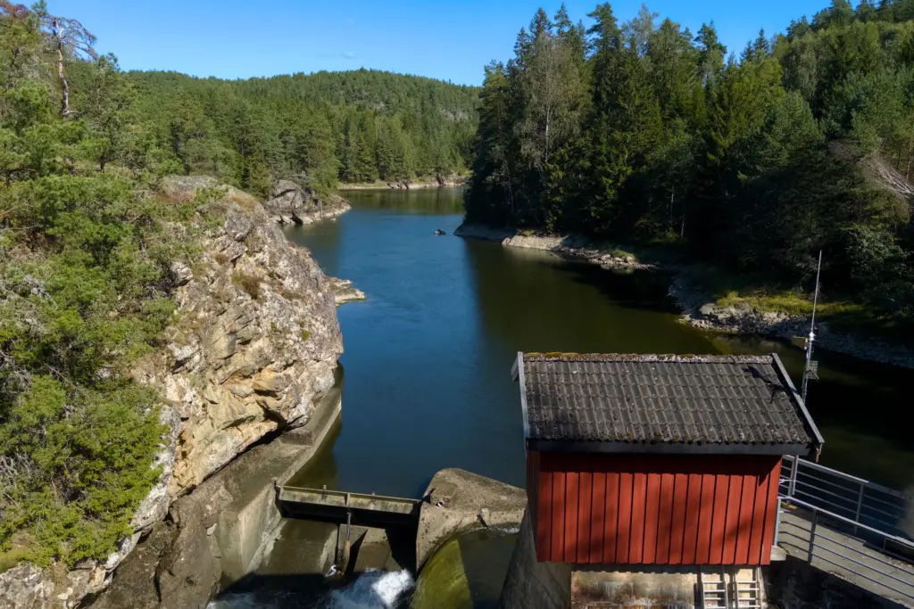 Liten vattenkraftsdamm med ett rött trähus vid en älv i barrskog en solig dag, en bild för jämförelsen av elavtalen från Vattenfall och E.ON