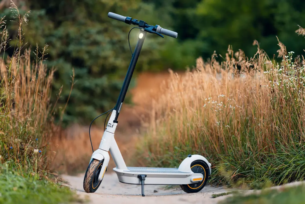 Vit elsparkcykel parkerad på en grusväg mellan högt gräs och granar en sommarkväll