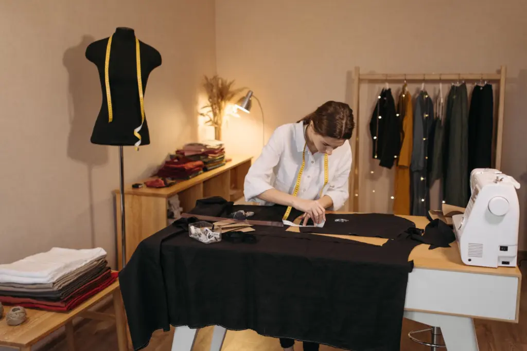 Sömmerska med måttband runt halsen ritar mönster på svart tyg i sin ateljé, med provdocka, klädställning och symaskin, en bild för att välja företagsform