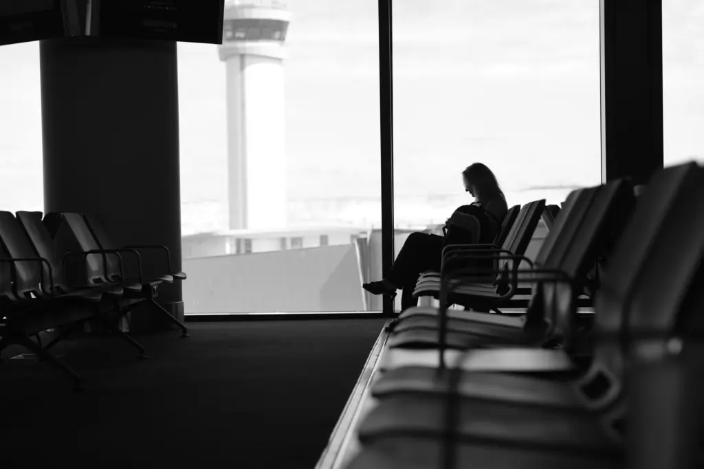 Svartvit bild av en resenär som väntar ensam i en flygplatsterminal med flygledartornet utanför fönstret