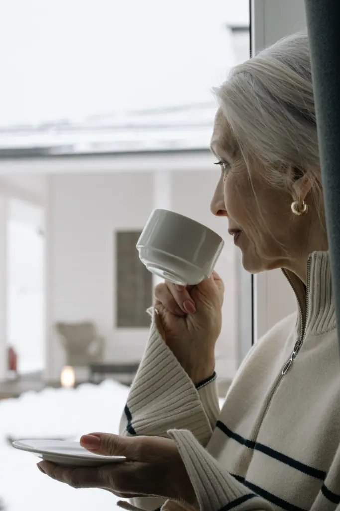 Äldre kvinna dricker kaffe vid fönstret en vinterdag, om garantipensionen för den som haft låg eller ingen inkomst