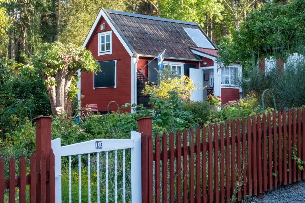 Röd trästuga med vita knutar och en solcellspanel på taket, bakom ett rött staket och en grönskande trädgård