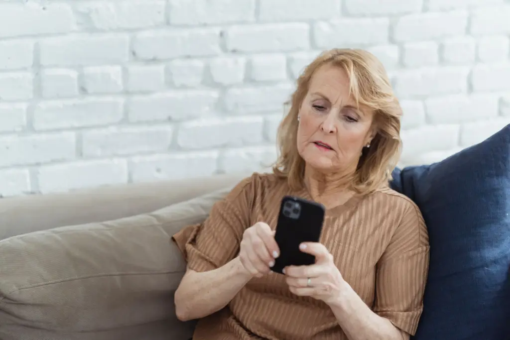 Kvinna i sextioårsåldern sitter i en soffa och tittar fundersamt på sin mobil