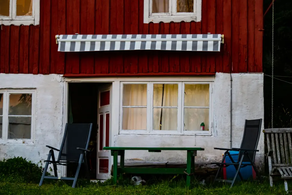 Röd stuga med vita knutar, markis och trädgårdsmöbler på gräsmattan, redo för sommarens hyresgäster