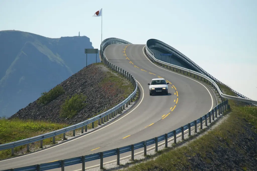 Vit bil kör över en välvd bro på en kustväg i Norge med berg i bakgrunden