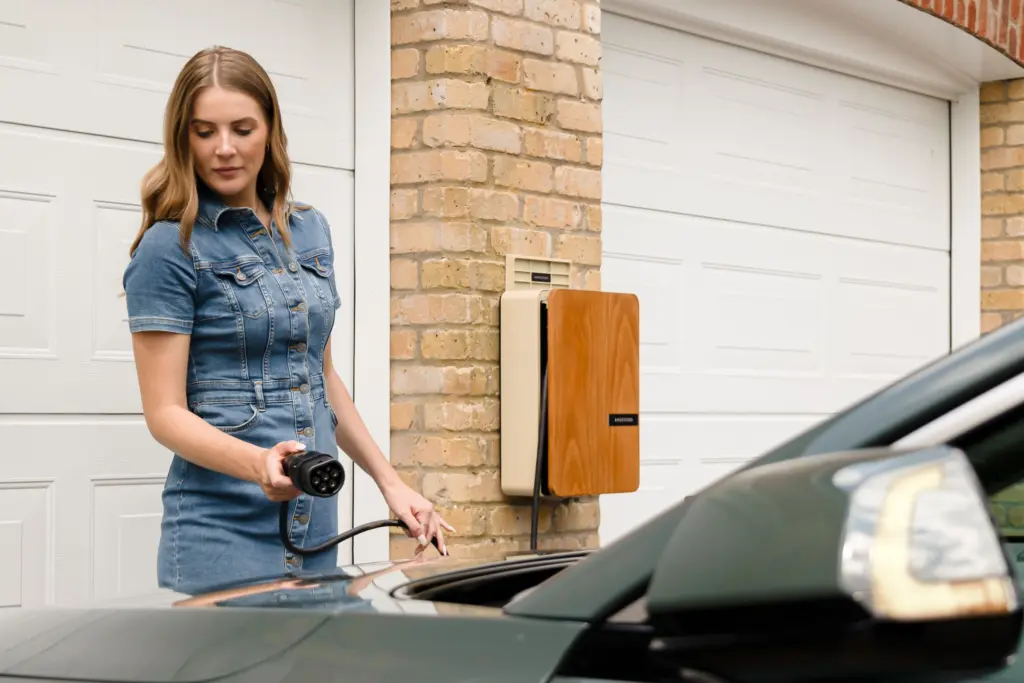 Kvinna kopplar laddkabeln från en väggmonterad laddbox vid garaget till sin elbil på uppfarten