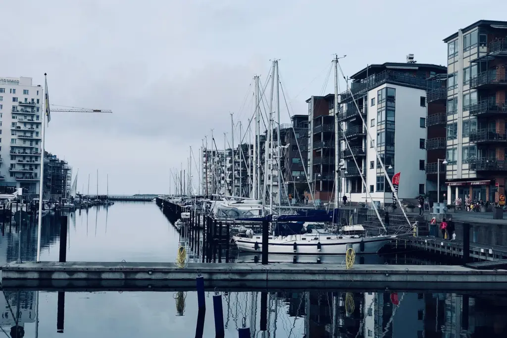 Segelbåtar förtöjda längs en brygga i en stadshamn i Skåne, med bostadshus vid kajen en mulen dag