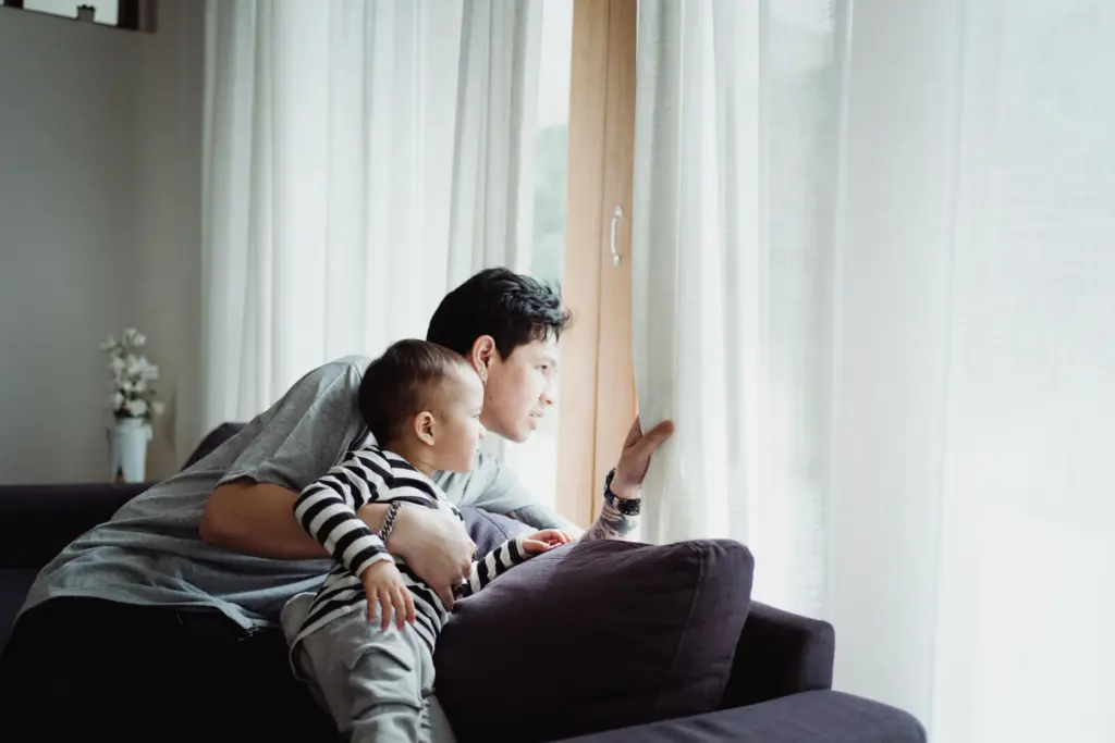 Pappa och litet barn i randig tröja tittar ut genom fönstret från soffan i ett ljust vardagsrum