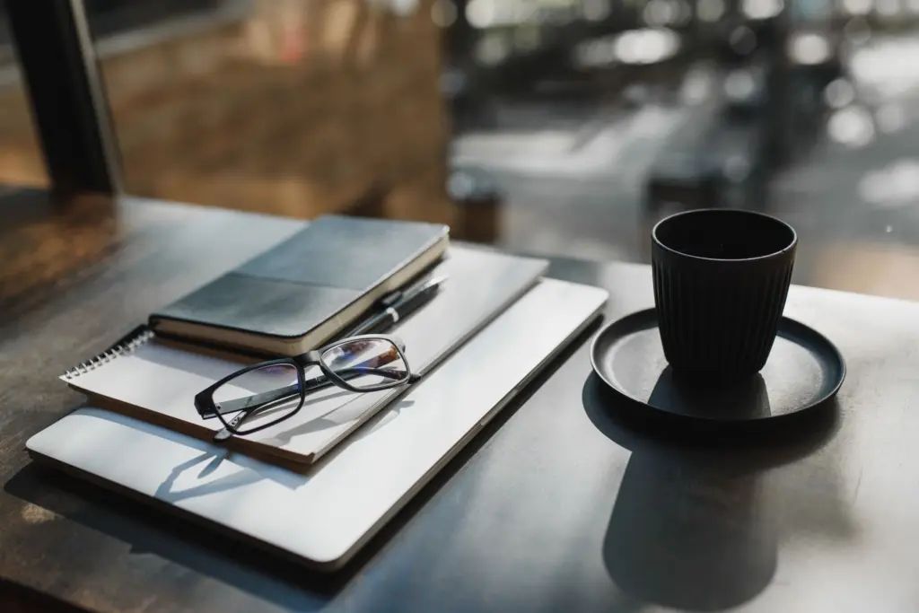 Anteckningsböcker, glasögon och en svart kaffekopp på ett mörkt bord vid ett fönster