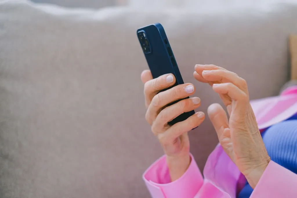 Äldre händer som håller och scrollar på en mobiltelefon i soffan, val av mobilabonnemang för pensionärer