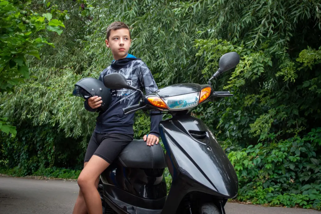 Tonårskille med hjälmen i handen står vid sin svarta moped på en väg kantad av grönska