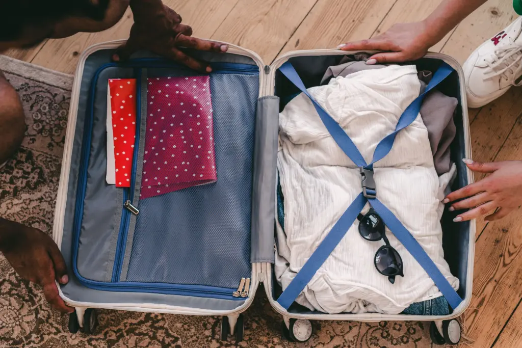 Två personer packar en öppen resväska med kläder, solglasögon och resedokument inför resan