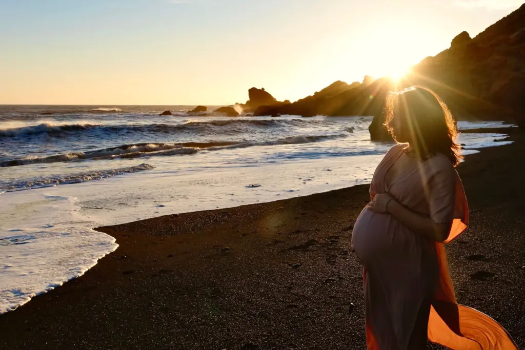 Gravid kvinna står på en svart sandstrand och ser ut över havet i motljus