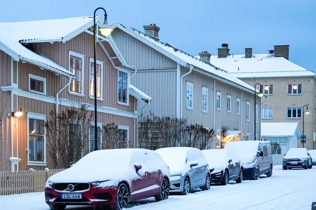 Snötäckta bilar parkerade längs en gata med trähus en vintermorgon, som när bilen står still och kan ställas av