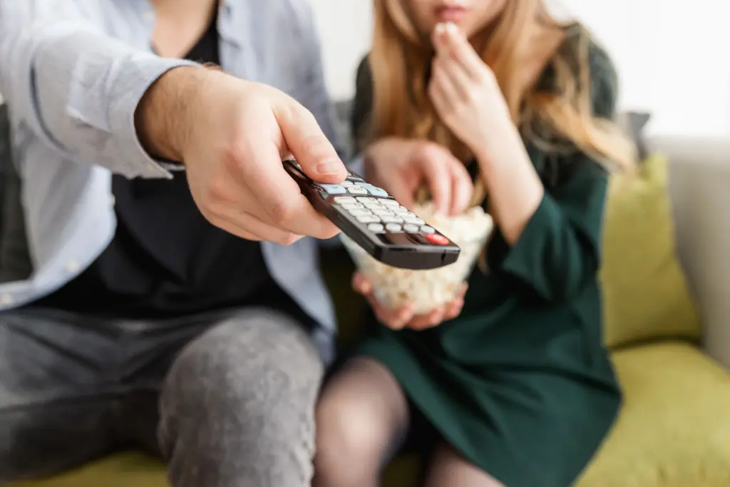 Ett par sitter i en soffa med en skål popcorn, och den ena riktar en fjärrkontroll mot tv:n