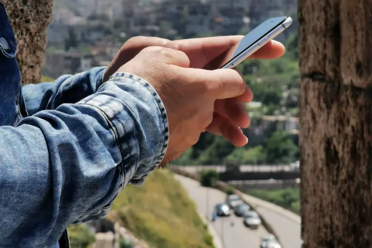 Hand som använder en mobiltelefon vid en stenmur med utsikt över en stad utomlands