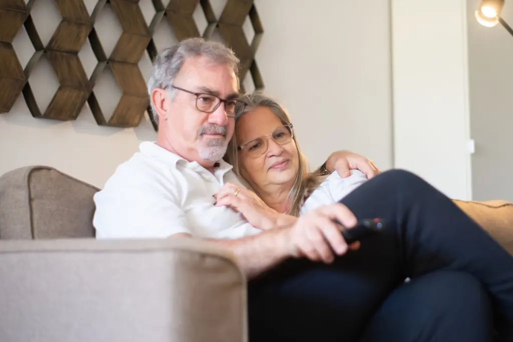 Äldre par sitter tätt ihop i soffan och tittar på tv med fjärrkontrollen i handen