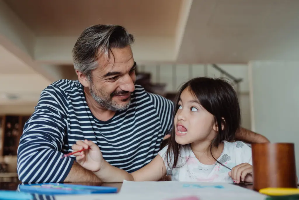 Pappa i randig tröja sitter vid köksbordet och skämtar med sin dotter som ritar, en bild för underhållet till barn när föräldrarna inte bor ihop
