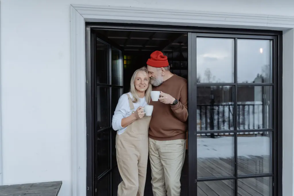 Äldre par med kaffekoppar står tätt intill varandra i dörröppningen till ett hus en vinterdag