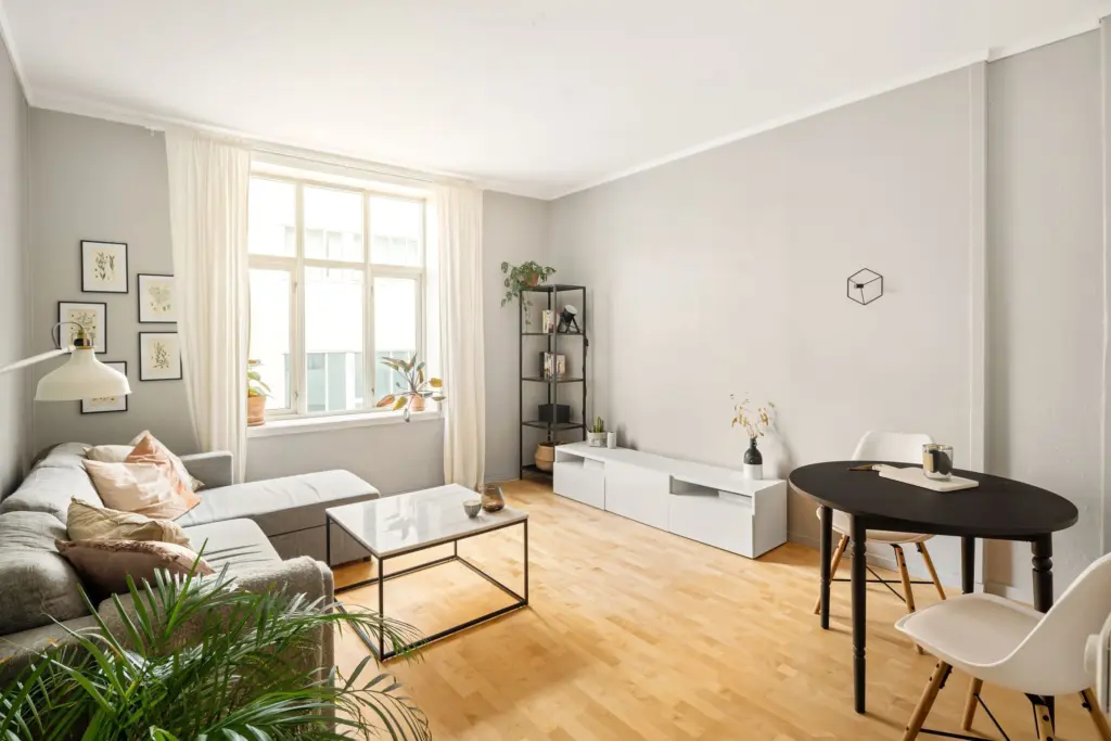 Ljust vardagsrum i en lägenhet med grå soffa, parkettgolv, högt fönster och ett litet matbord