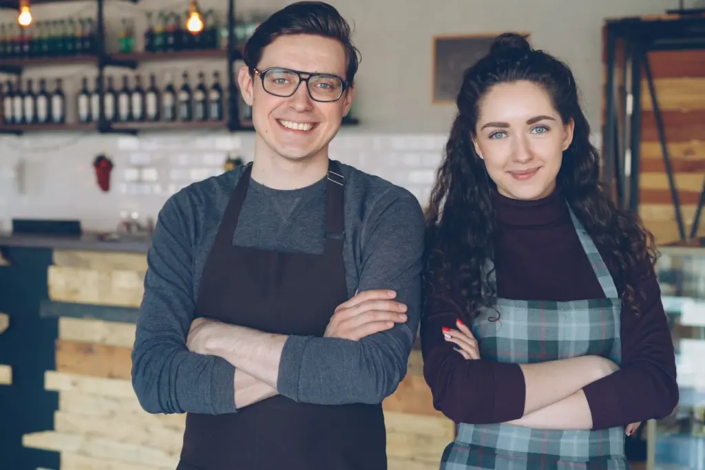 Två kaféägare i förkläden står med armarna i kors framför disken i sitt kafé