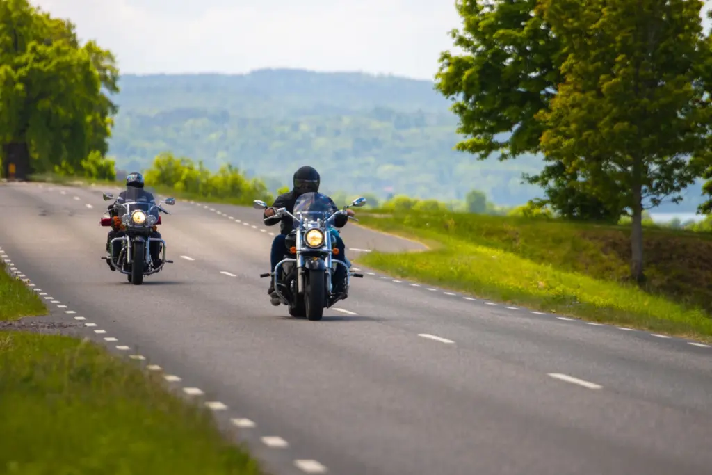 Två motorcyklister kör på en svensk landsväg en sommardag, med grönska och skogsklädda höjder i bakgrunden