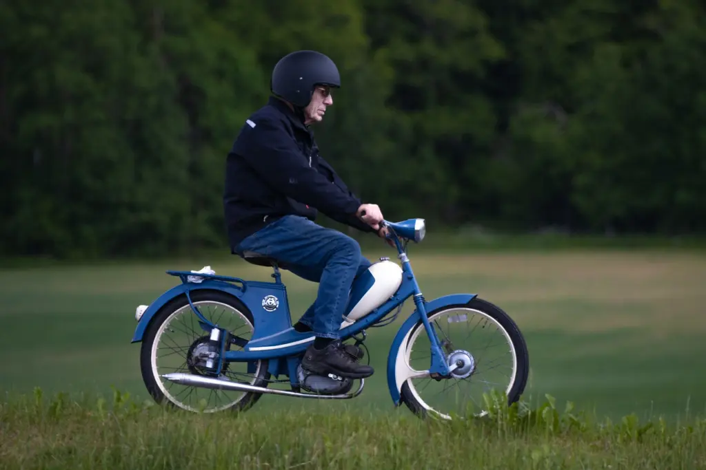 Man med hjälm kör en äldre blå moped över en gräsäng på den svenska landsbygden