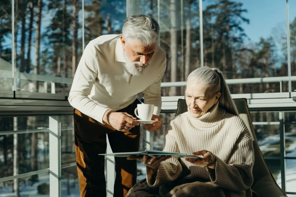 Äldre par med kaffe vid ett fönster mot snöig tallskog, en lugn vardag när pensionen ska jämföras och planeras