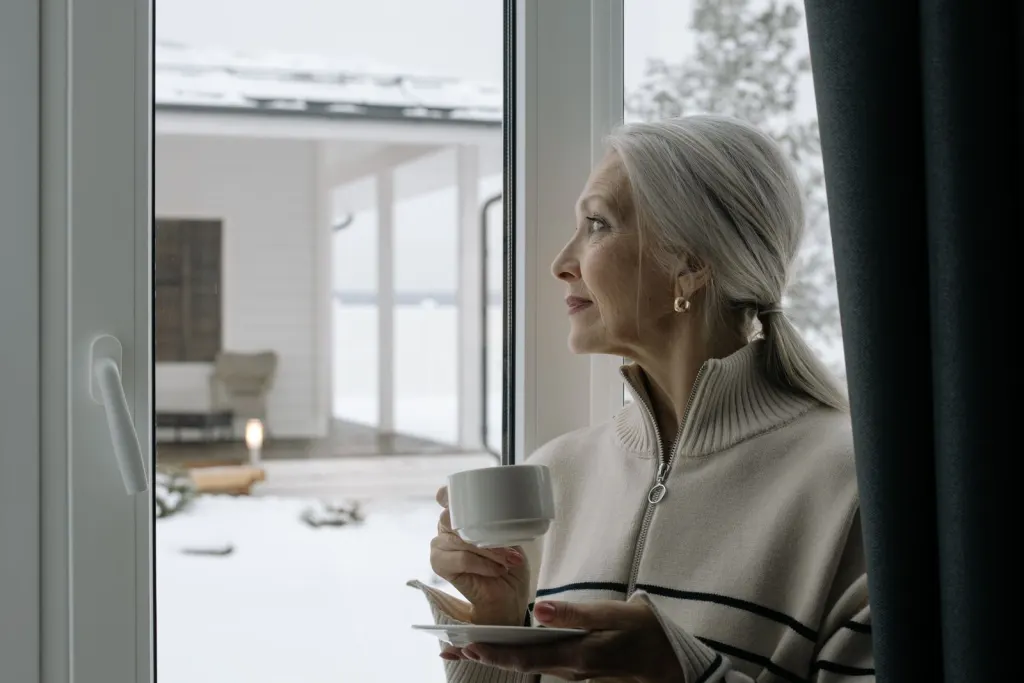 Äldre kvinna med kaffekopp tittar ut genom fönstret mot snön, trygg med pengar på sparkonto