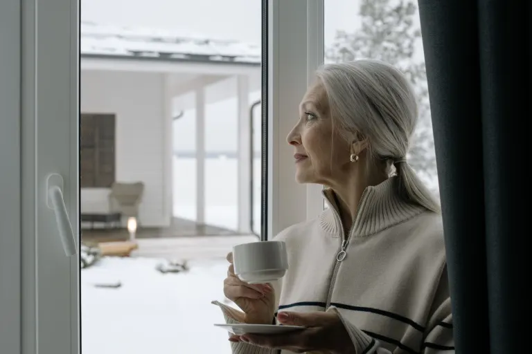 Äldre kvinna med kaffekopp tittar ut genom fönstret mot snön, trygg med pengar på sparkonto